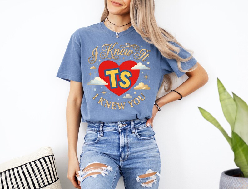 May include: A blue short-sleeve t-shirt featuring a red heart graphic with the yellow text TS inside. The design includes gold cursive text reading I Knew It and I Knew You, surrounded by small white clouds and gold stars printed on the front.
