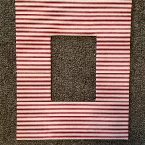 May include: A rectangular fabric frame with a square cutout in the center. The fabric features a pattern of thin, horizontal red stripes on a white background. The frame is likely for displaying photos or artwork.