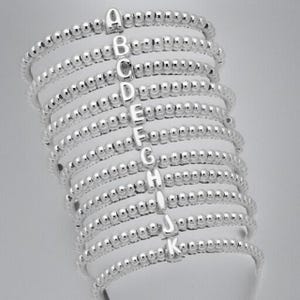 May include: A stack of silver beaded bracelets, each featuring a different letter from A to K. The bracelets are made of small, round beads and are displayed on a white surface against a light gray background. The letters are in a simple, sans-serif font.