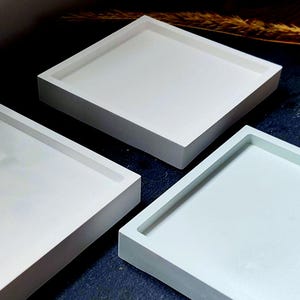 Könnte beinhalten: Drei quadratische, minimalistische Tabletts in Weiß- und Hellblautönen sind auf einer dunkelblauen Oberfläche angeordnet. Die Tabletts haben ein schlichtes, modernes Design mit leicht erhöhtem Rand. Ein Zweig getrocknetes Gras setzt einen natürlichen Akzent.