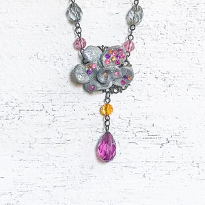 Op de afbeelding: Een zilverkleurige bloemenhangerketting met roze, oranje en paarse accenten. De ketting heeft een bloemvormige hanger met ingebedde strass-steentjes en een bungelende druppelvormige bedel. De ketting is versierd met grijze en roze kralen.