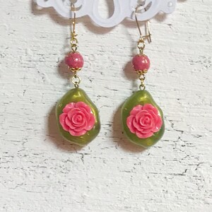 Op de afbeelding: Een paar oorhangers met goudkleurige haken, roze kralen en olijfgroene druppelvormige hangers. Elke hanger heeft een verhoogd roze rozenontwerp. De oorbellen hangen tegen een witte achtergrond.