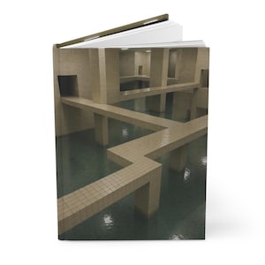 May include: Hardback journal with a surreal architectural design on the cover. The image shows a beige tiled structure with walkways and water, creating a maze-like appearance. The open pages are visible.