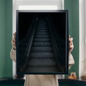 May include: A framed print featuring a dark, dramatic image of an escalator ascending into darkness. The steps are a dark gray, with subtle sparkling highlights. The frame is black, creating a stark contrast.