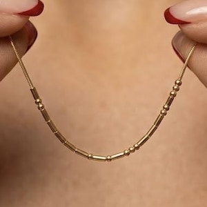 May include: A gold-toned necklace featuring a delicate chain adorned with a series of small, cylindrical beads and round spheres arranged in a specific pattern, creating a linear design that rests against the skin in a gentle, curved arc.
