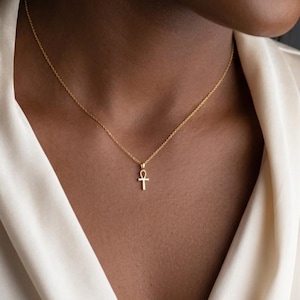 May include: A delicate gold necklace featuring a small ankh pendant suspended from a thin, polished chain. The pendant is shaped like a cross with a loop at the top, centered on the collarbone area against a smooth, light-colored fabric.