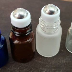 May include: Four small glass roller bottles with silver rollerballs and white caps. The bottles are in various colours: blue, brown, frosted, and clear. These bottles are suitable for essential oils, perfumes, or other cosmetic products.