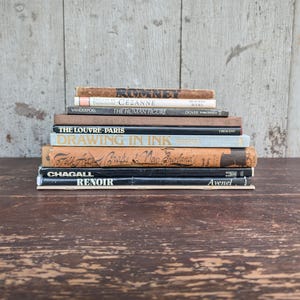 May include: A stack of vintage books with various titles, including "Cezanne," "The Human Figure," and "Drawing in Ink." The books are in different colors, with aged covers and visible wear, resting on a dark wooden surface.
