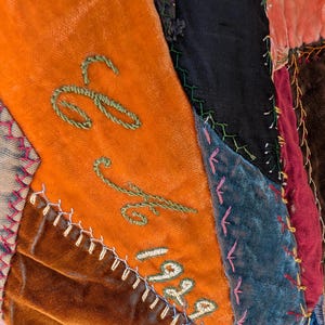 May include: Close-up of a patchwork textile with various velvet fabrics in orange, brown, blue, and black. The orange section features green embroidered cursive letters and the number "1929". The edges are stitched with white thread.