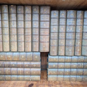 May include: A collection of antique books with aged leather covers, arranged in a wooden tray. The spines display titles and decorative gold-colored accents. The books are in varying shades of tan and brown.