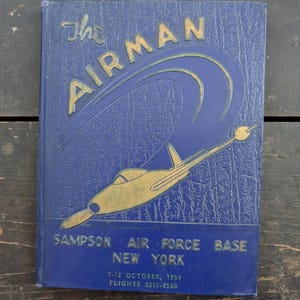 May include: A vintage blue book titled "The Airman" with gold lettering and a gold airplane illustration. The book cover also includes the text "Sampson Air Force Base New York" and the date "7-12 October, 1954".
