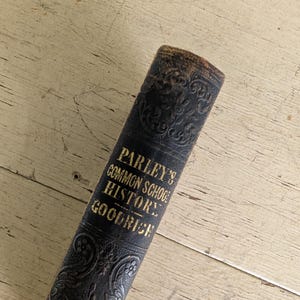 May include: A dark brown, antique book spine with embossed floral patterns and gold lettering that reads Parley's Common School History Goodrich. The spine shows signs of age and wear, with a textured surface and visible decorative detailing.