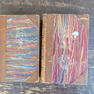 May include: Two antique hardcover books featuring brown leather spines and corners with marbled paper covers. The marbled patterns display intricate wavy lines in red, blue, yellow, and white. The spines show raised bands and signs of wear.