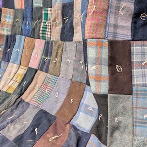 May include: A patchwork quilt constructed from rectangular fabric pieces in various shades of blue, gray, brown, and green. The textile features a mix of solid colors and plaid patterns, with small, light-colored thread knots visible on each square.