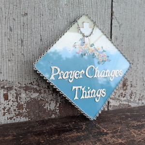 May include: A diamond-shaped wall hanging with a blue and white gradient background. It features a white cross, a cluster of small colorful flowers, and the white text Prayer Changes Things. The piece is framed with a silver-toned metal chain border.