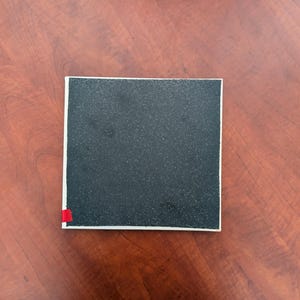 May include: A square, black object with a textured surface and a white border. A small, red accent is visible on one corner. The object is set against a warm, reddish-brown wooden surface.