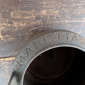 May include: An antique, cast iron pot with the word "MARIETTA" embossed on the rim. The pot has a dark, weathered appearance, with signs of age and use. The background is a distressed wooden surface.