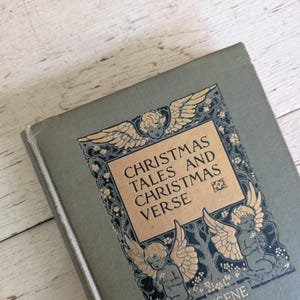 May include: A hardcover book titled Christmas Tales and Christmas Verse by Eugene Field with a sage green cover. The front features a gold rectangular panel with black text, surrounded by dark blue and gold illustrations of three winged cherubs.