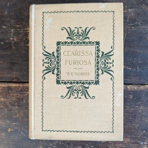 May include: A vintage book titled "Clarissa Furiosa" by W.E. Norris. The book has a beige cover with green decorative elements and text. The cover design includes a central square with the title and author's name, framed by ornate flourishes.