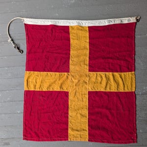 May include: A red and yellow nautical flag with a white border and rope. The flag features a yellow cross on a red background. The text "INT N 6" is printed on the white border.