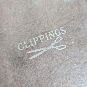 May include: A textured, taupe-colored surface featuring the word CLIPPINGS printed in a light, sans-serif font, accompanied by a graphic illustration of a pair of open scissors positioned directly beneath the text in a matching light tone.