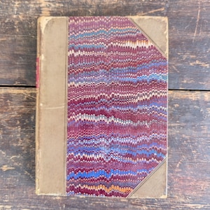 May include: Antique book with a marbled cover in shades of burgundy, blue, and gold. The book has a worn, tan spine and corners, suggesting age and use. The cover design is a classic example of decorative bookbinding.