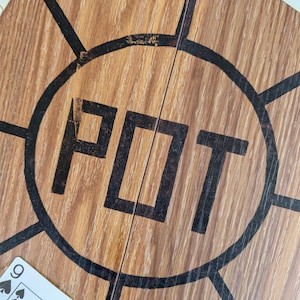 May include: Close-up of a wooden poker table surface with the word "POT" in large black letters inside a black circle. Playing cards with visible suits and numbers are scattered around the table, suggesting a game in progress.