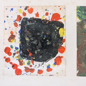 May include: Abstract artwork featuring a black square with colorful paint splatters in red, yellow, and blue on a white background. The artwork is framed in a white frame. The style is reminiscent of abstract expressionism.