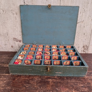 May include: An open, vintage blue wooden box with multiple compartments, each labeled with letters and numbers. The box has a hinged lid and a metal clasp. Some compartments contain small, decorative items.