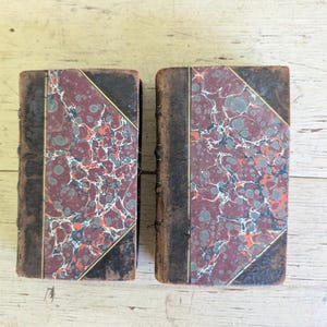 May include: Two antique books with rectangular covers featuring a marbled pattern in shades of deep red, gray, and white. The covers are accented with dark brown leather edges and thin gold-colored lines, showcasing a classic bookbinding style.