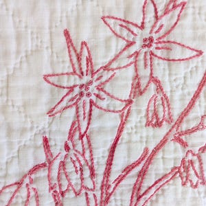May include: Close-up of a white quilted fabric with hand-stitched embroidery. The embroidery features a floral design in shades of pink, with detailed flower and stem outlines. The stitching is visible and adds texture to the fabric.