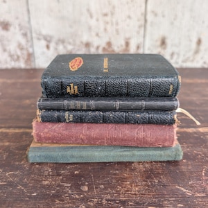 May include: A stack of four antique books with worn covers. The top book is black with gold lettering and a red emblem. The other books have covers in shades of black, red, and teal. The books are stacked on a wooden surface.