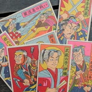 May include: A collection of vintage Japanese trading cards featuring illustrations of samurai warriors in various action poses. The cards have vibrant colors and Japanese text, depicting scenes of combat and historical figures.