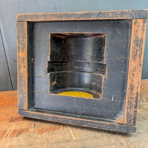 May include: A square wooden mold with a distressed black and natural wood finish. The center features a deep, cylindrical cutout with smooth black interior walls and a bright yellow circular base visible at the bottom of the cavity.