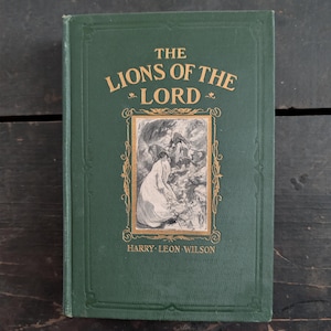 May include: A vintage hardcover book titled "The Lions of the Lord" by Harry Leon Wilson. The cover is dark green with gold lettering and a framed illustration. The book is in good condition, showing signs of age.