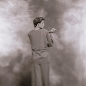 May include: Sepia-toned photograph of a person wearing a long-sleeved, belted dress and pants. The person is turned away from the camera, with their head in profile. The background is a blurred, cloudy effect.