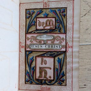 May include: An illuminated manuscript page featuring the text Imitation of Jesus-Christ. The design includes blue and gold floral motifs, a central IHS symbol, and the word Caritas at the bottom, all framed by intricate red ink line work on paper.