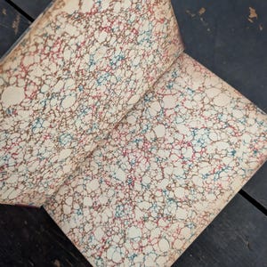 May include: Open book with marbled endpapers. The paper has a cream background with a pattern of red, blue, and brown swirls. The book's spine is visible, with a maroon color. The pages are slightly aged.