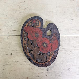 May include: A dark brown wooden artist palette featuring a carved and painted floral design. The surface displays three prominent red poppy flowers with detailed black centers, surrounded by intricate dark wood grain textures and patterns.