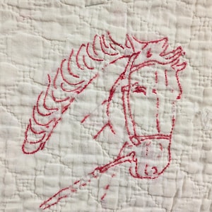 May include: White quilted fabric with a red embroidered horse head design. The horse head is detailed with a bridle and reins, showcasing a classic equestrian theme. The stitching is visible, adding a handcrafted feel.