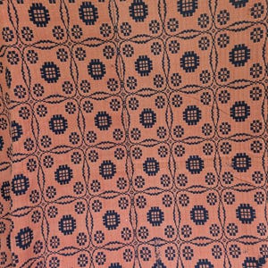 May include: Close-up of a textile with a repeating pattern. The fabric has a peach-colored background with a dark blue geometric design. The pattern includes squares and circles with cross-hatch details, creating a textured appearance.