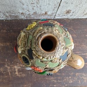 May include: A round ceramic jug with a textured surface covered in a mosaic of various colorful plastic buttons, beads, and small decorative shapes in shades of red, green, yellow, and black, featuring a single handle and a central opening.