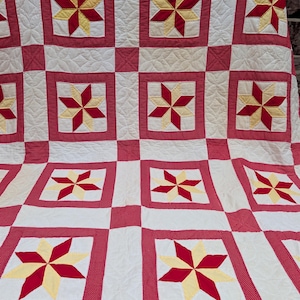 May include: A quilt featuring a repeating grid pattern of red and white squares. Each white square contains a six-pointed star motif composed of alternating red and pale yellow diamond-shaped fabric pieces. The red squares feature a fine white dot pattern.