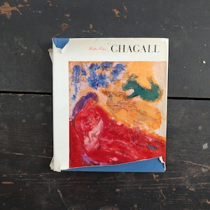May include: A book cover featuring the name "CHAGALL" in black capital letters. The artwork depicts a figure in red, with blue and yellow accents. The cover has a white border with some wear and tear.