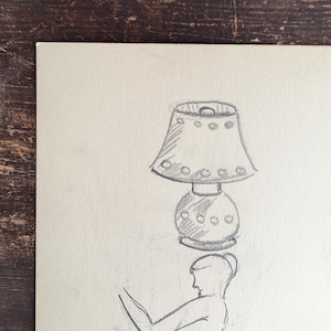 May include: A graphite pencil sketch on off-white paper depicting a figure balancing a table lamp on their head. The lamp features a wide shade with circular decorative accents and a rounded base. The figure is shown in profile with arms raised.