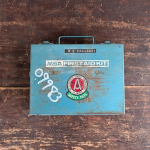 May include: A vintage, light blue metal first aid kit with a handle. The kit has the text "MSA FIRST AID KIT" and a circular emblem with the text "SAFETY PAYS". The number "09983" is written on the front.