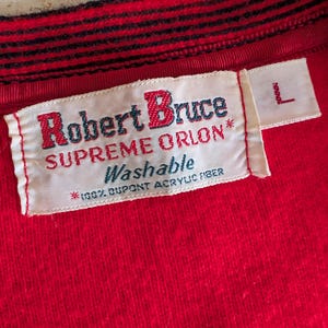 May include: A bright red garment featuring a white rectangular clothing label with the text Robert Bruce Supreme Orlon Washable 100% Dupont Acrylic Fiber and a separate small white tag with the letter L. The collar has black and red stripes.