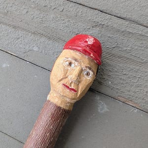 May include: Hand-carved wooden figure with a painted face and a red cap. The figure has a brown, textured stem. The face is painted with brown eyes and red lips. The figure is likely a decorative item or folk art.