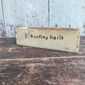 May include: A rectangular, weathered, light-colored wooden storage box with open top. The front panel features the black hand-painted text "3/4 Roofing nails". The wood shows signs of age, including surface wear, discoloration, and visible grain.