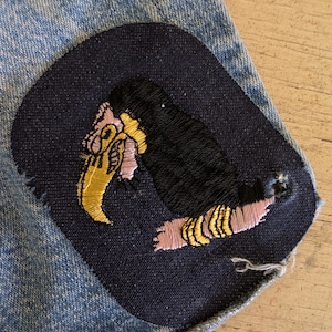 May include: A dark blue fabric patch featuring an embroidered toucan with a yellow beak, black body, and pink accents, stitched onto a light blue denim garment. The patch has a rounded rectangular shape with visible thread details throughout.